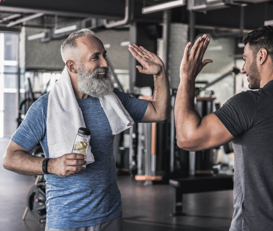 Natural Pharmaceuticals Vitamiin K2 Complex Kaks meest jõusaalis tervitavad üksteist käepigistusega, üks hoiab veepudelit ja rätikut.